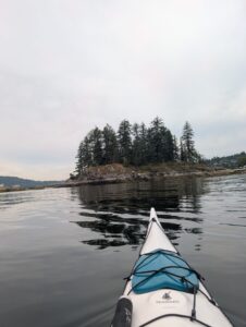 Kayak bow pointing toward a forested rocky island in calm coastal waters, with evergreen trees and distant shoreline visible under overcast sky